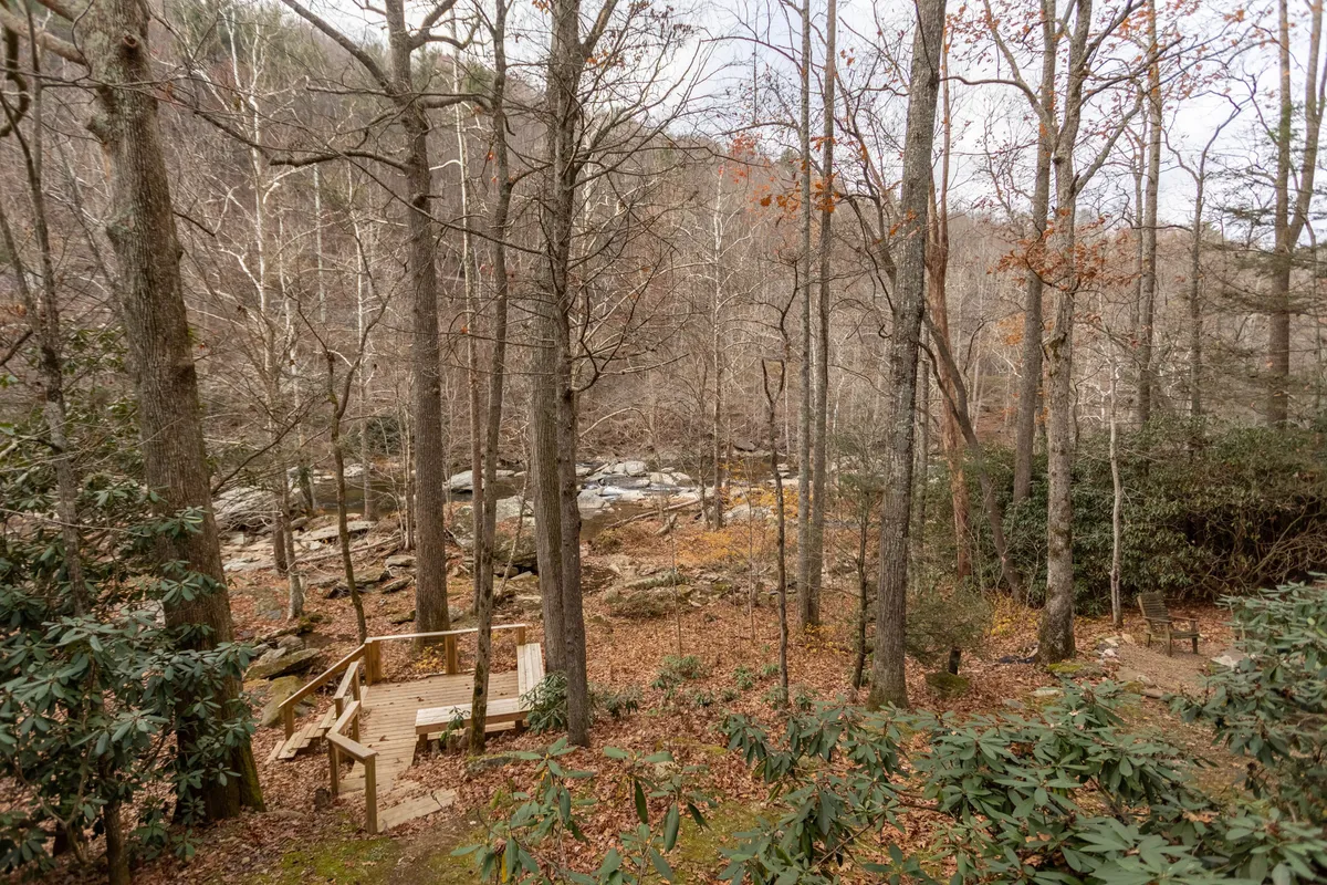 A spacious wooden deck overlooks a rocky stream framed by bare trees and green shrubs. The tranquil riverside setting is perfect for enjoying fresh air and nature, with easy access to the water's edge for walks or playtime.