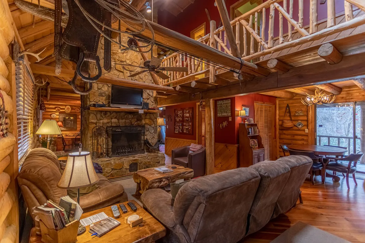 This image captures a spacious great room featuring a dramatic stone fireplace flanked by comfortable seating and wooden architectural beams overhead. The dining area is visible in the background, emphasizing the room's open concept and social layout. Large windows likely offer glimpses of the surrounding natural scenery, contributing to a bright, inviting atmosphere.