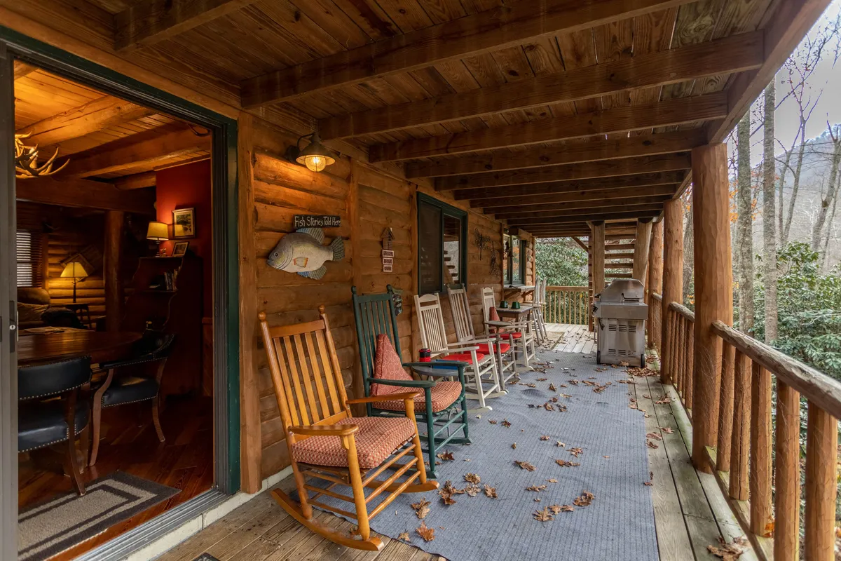 A cozy exterior scene with rocking chairs lined up on a wooden deck, a grill, and wooden railing. The chairs face outwards, inviting guests to relax, watch sunsets, or share stories while overlooking the river and enjoying the fresh mountain air.