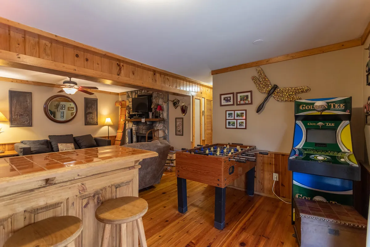A vibrant game room with a foosball table, arcade machine, bar area, and comfortable seating. The space is decorated with playful wall art, and natural light brightens the room. The blend of entertainment features and a relaxed seating area invites both family and friends to enjoy game nights or casual lounging.