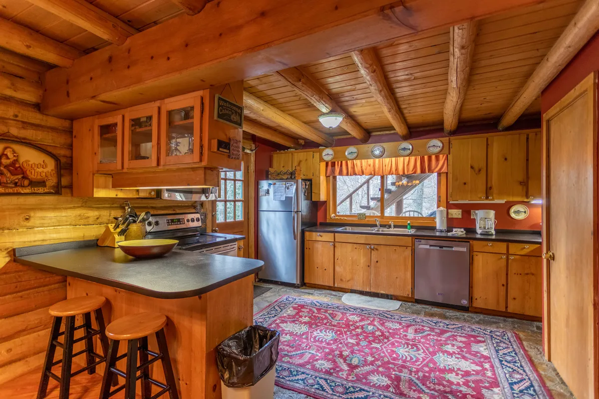 A cozy, well-lit kitchen with wooden cabinets, a spacious countertop, a large window overlooking nature, and bar stools tucked under the counter. The rug along the floor adds warmth, and the scene invites both meal preparation and casual conversation. The layout and natural light exemplify a welcoming space for cooking and gathering.