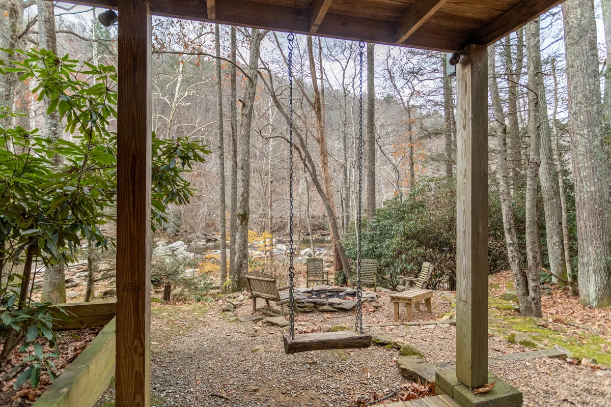 A scenic outdoor area by the river with a swing, fire pit, rustic wooden seating, and lush trees. The setting invites riverside gatherings, storytelling by the fire, and relaxing while listening to the river, offering activities for all ages.