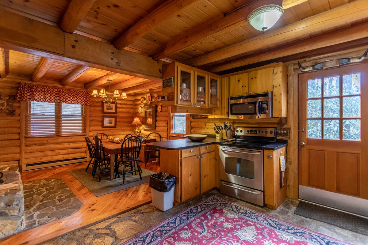 A wide-angle view of the dining area reveals a sturdy wooden table with seating for six, set beside a striking stone fireplace. The scene includes warm wooden cabinetry and floor rugs, with kitchen elements nearby, reflecting a communal space for meals and togetherness.