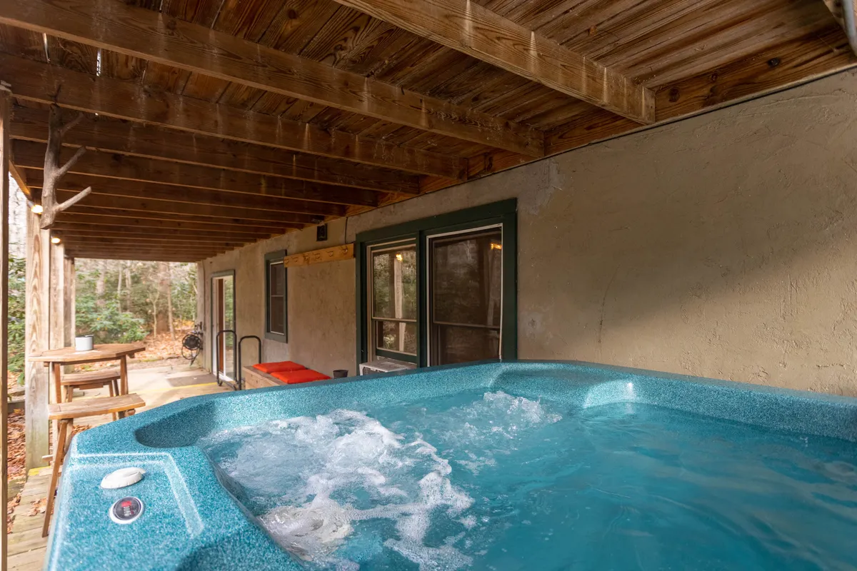 A tranquil back porch features a hot tub under a wooden ceiling, surrounded by views of trees. The inviting setup suggests relaxation after outdoor adventures, with hints of both privacy and immersion in nature.