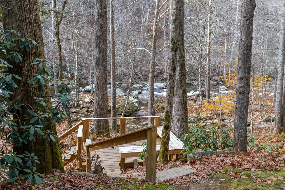 A peaceful wooden deck overlooks the flowing river, with autumn leaves scattered among bare trees. The deck is a perfect spot for bird watching, reading, or simply soaking up the tranquil riverside atmosphere. The river is prominent, emphasizing the property’s immersive natural setting.