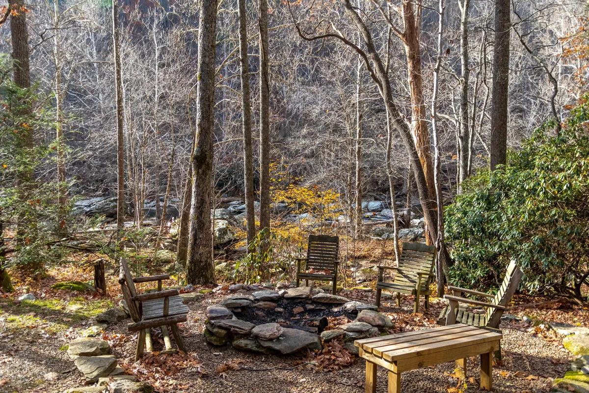 This image features a riverside fire pit surrounded by wooden chairs, set among natural rock formations and tall trees. The setting is ideal for campfire storytelling, stargazing, and group gatherings in a tranquil, riverside environment.