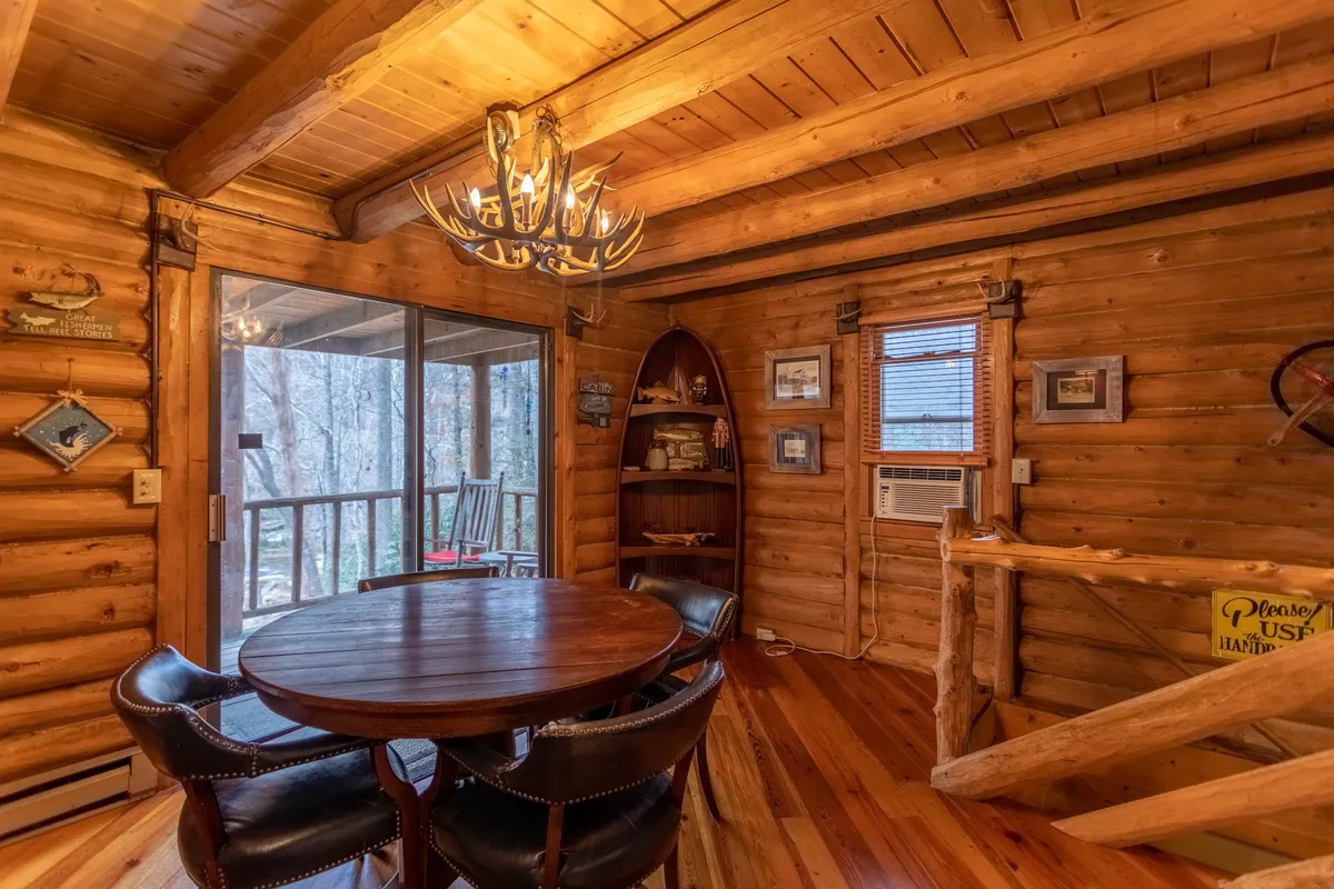 A warmly lit dining space features a large wooden table, elegant chandelier, and expansive windows that offer glimpses of the outdoors—likely the river view mentioned in the text. Wooden walls and decorative items create a welcoming environment, hinting at easy access to the deck.