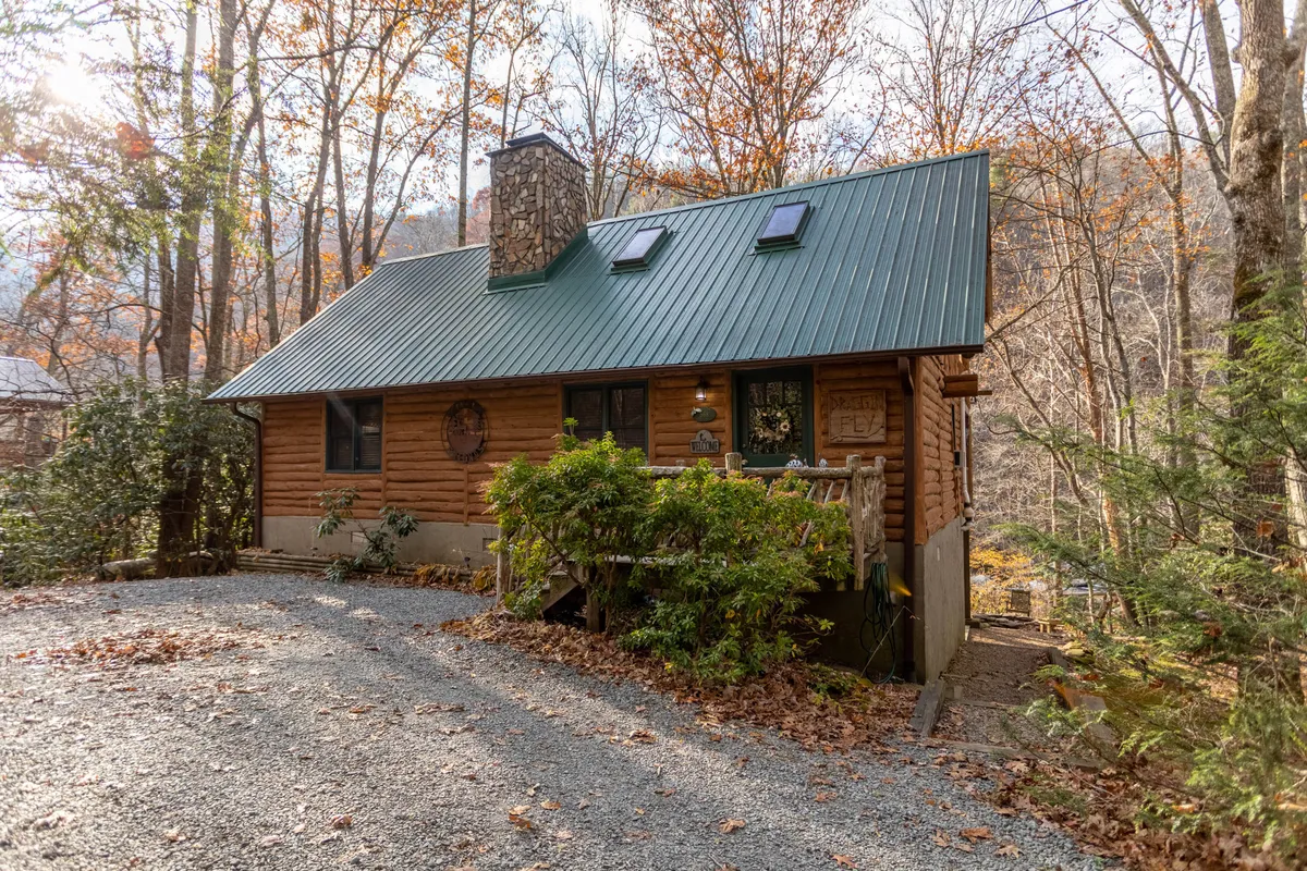 This image showcases the inviting exterior of a classic log cabin nestled among lush greenery, with a striking green metal roof, a rustic stone chimney, and a welcoming front porch complete with railings. The serene setting hints at peaceful moments spent watching sunsets, gathering outdoors, and enjoying the tranquil riverfront location.