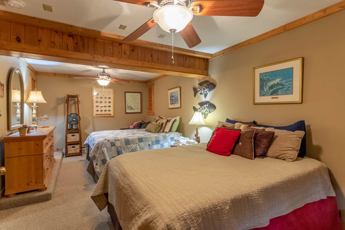 A spacious bedroom with two beds, a dresser, ceiling fan, and subtle wall decor. The room feels cozy and functional, suitable for families or friends. The color palette and furnishings create an inviting, restful environment.