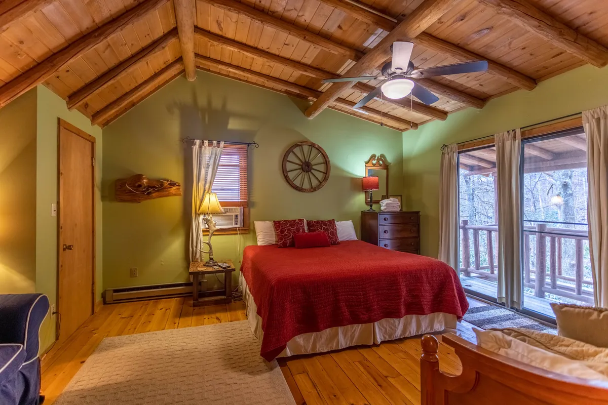 A warm bedroom featuring a bed with a red blanket, wooden ceiling, and a large window that opens to a balcony. A decorative wheel hangs on the wall, adding rustic charm. The room is filled with natural light and offers inviting views of nature.