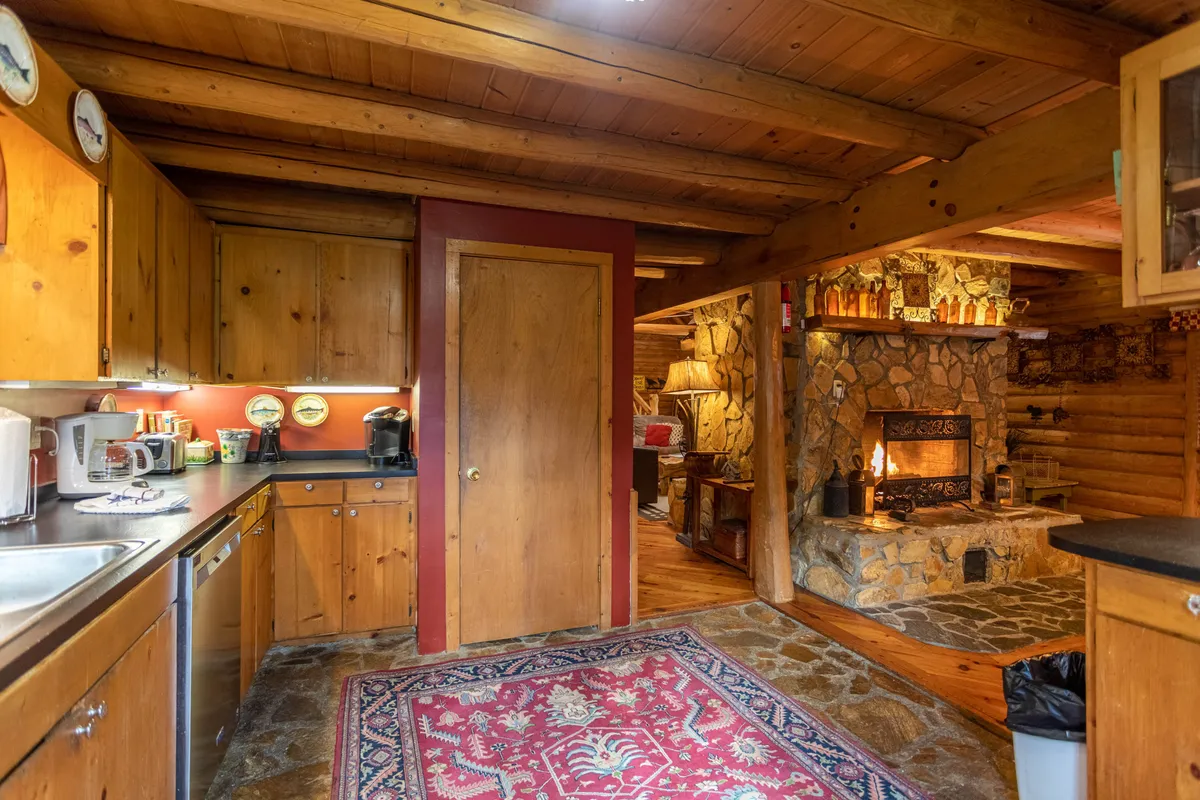 A charming kitchen and breakfast nook area featuring warm wooden cabinetry, a stone fireplace, and a colorful rug. The visible round table is positioned near a large window, perfect for enjoying coffee with a view of the water, while the cozy fireplace adds a welcoming touch for intimate meals.