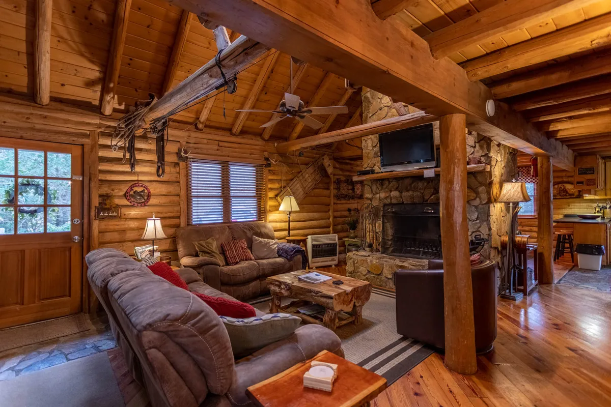 The image showcases a cozy seating area arranged around a robust stone fireplace, with exposed wooden beams overhead. Plush sofas and a coffee table invite relaxation, while the overall design exudes rustic charm. Natural light filtering through the windows enhances the bright, heartwarming feel.