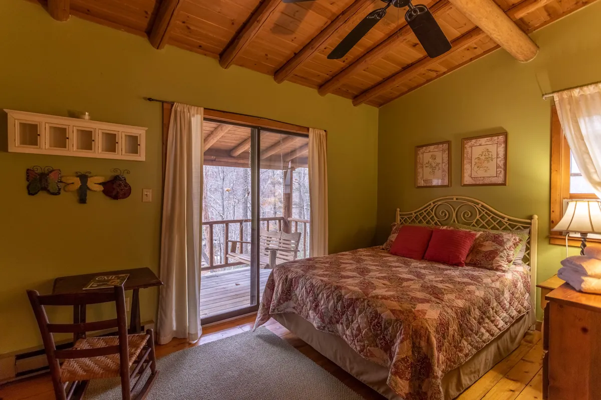 A cozy bedroom with a quilted bed, wooden ceiling, and a sliding glass door that leads to an upper deck. There is a small wooden table and chair near the door, offering a spot to enjoy views or write. Morning light streams in, with a sense of tranquility and comfort throughout the space.
