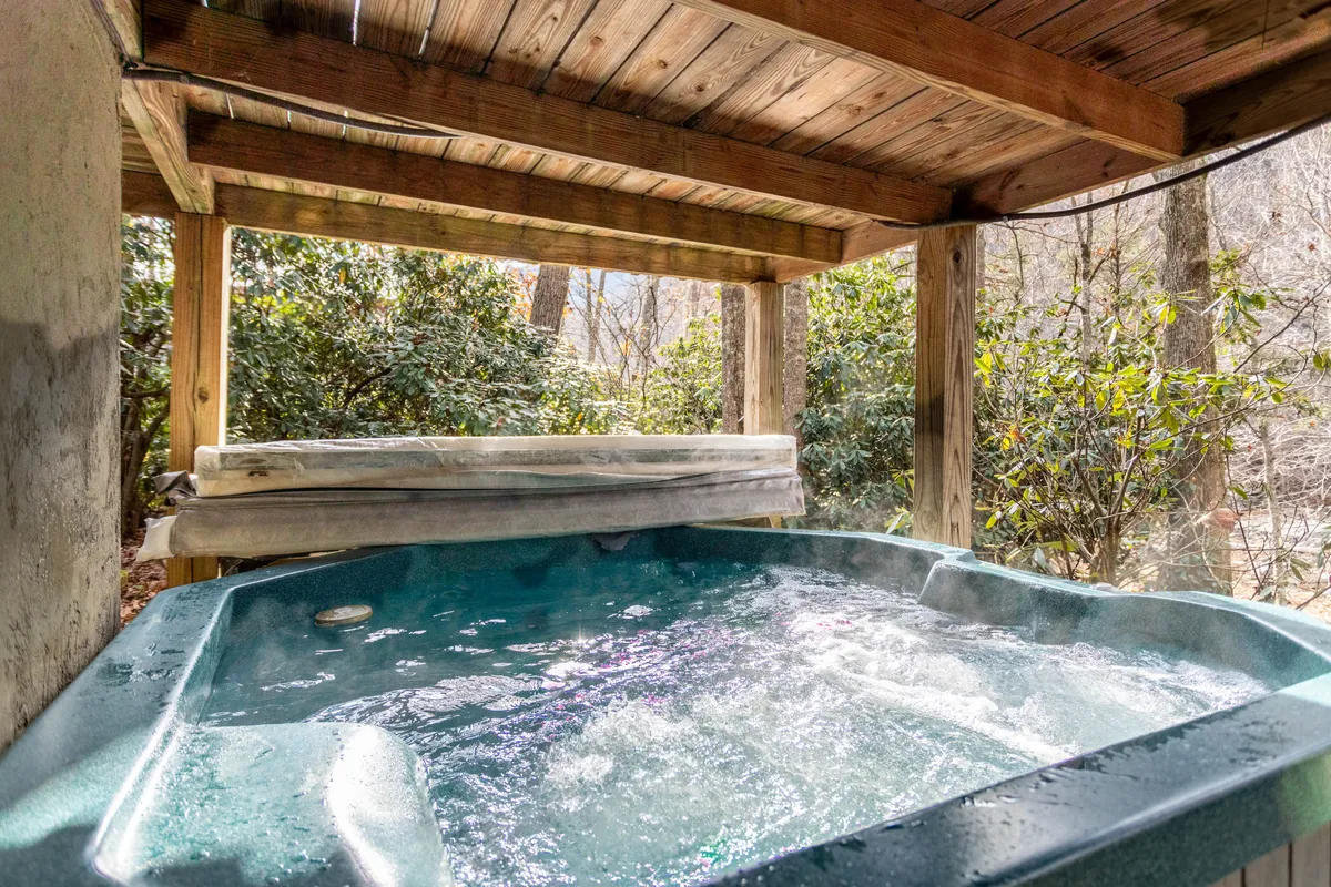 This image showcases a tranquil riverside hot tub surrounded by lush greenery and wooden beams. The hot tub sits on a deck overlooking the water, capturing the perfect blend of comfort and natural beauty, ideal for relaxing or stargazing in the evening.
